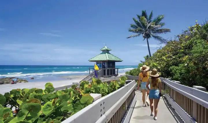 Two people in summer clothes and hats walk on a boardwalk toward a beach. A lifeguard tower with a green roof and palm tree are visible.