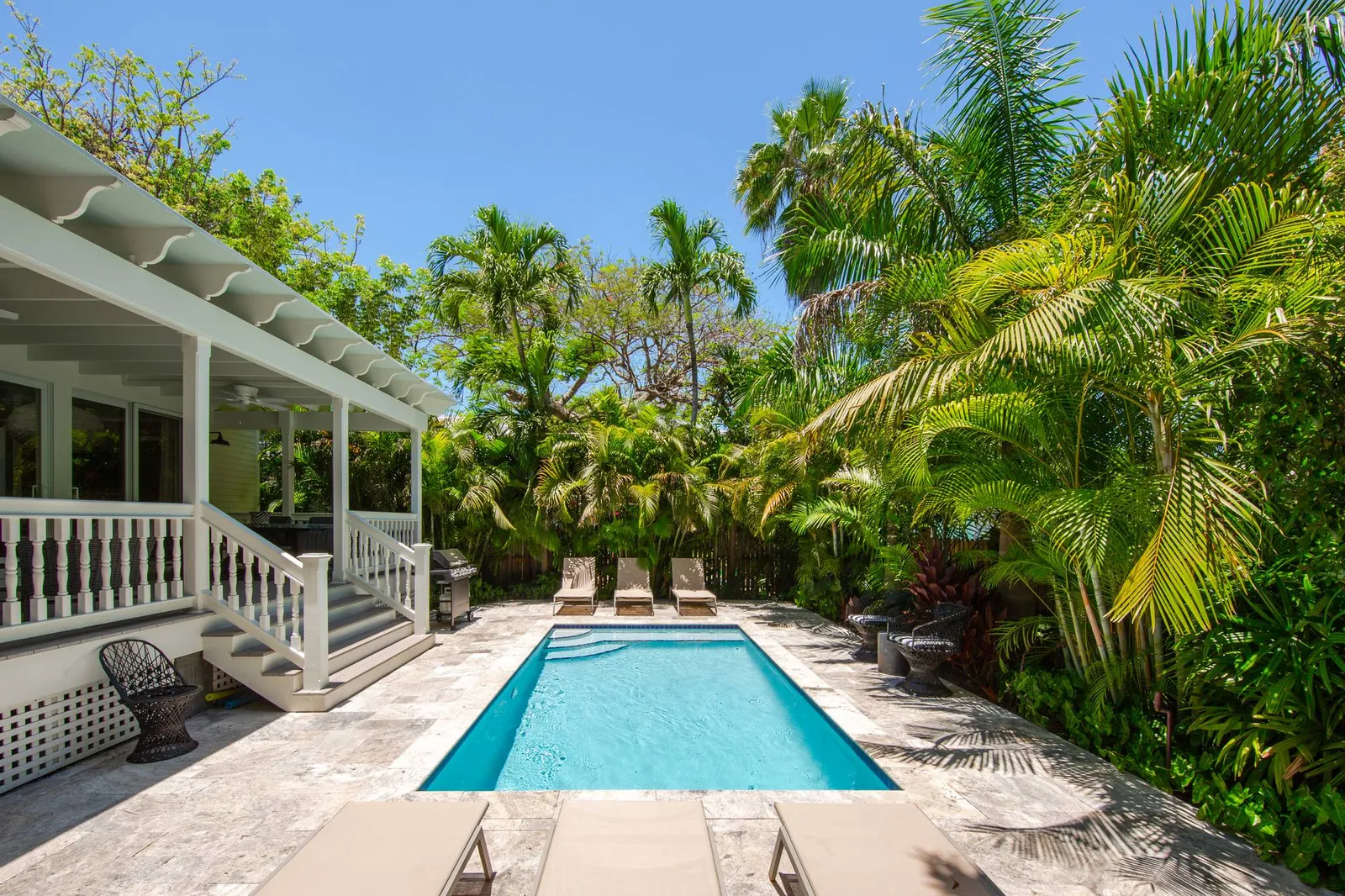 A backyard pool with lounge chairs surrounded by lush palm trees. A shaded patio with chairs and white railing is on the left. Sunny day.