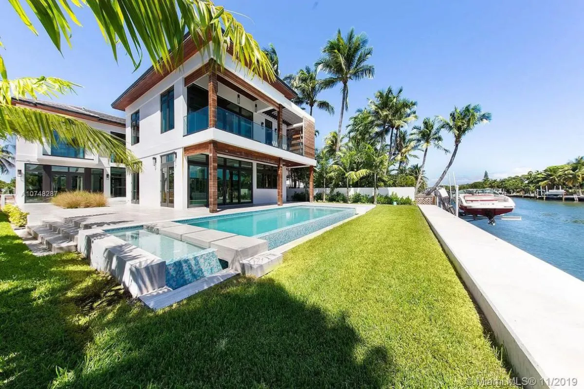 Modern white house with large windows, pool, and lush lawn. Palm trees surround, with a boat on a canal. Sunny, clear blue sky.